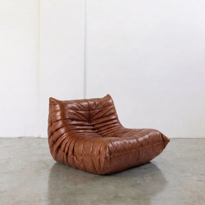 Togo Chair Dupe in brown real shiny oil wax leather on a concrete floor with a white wall background