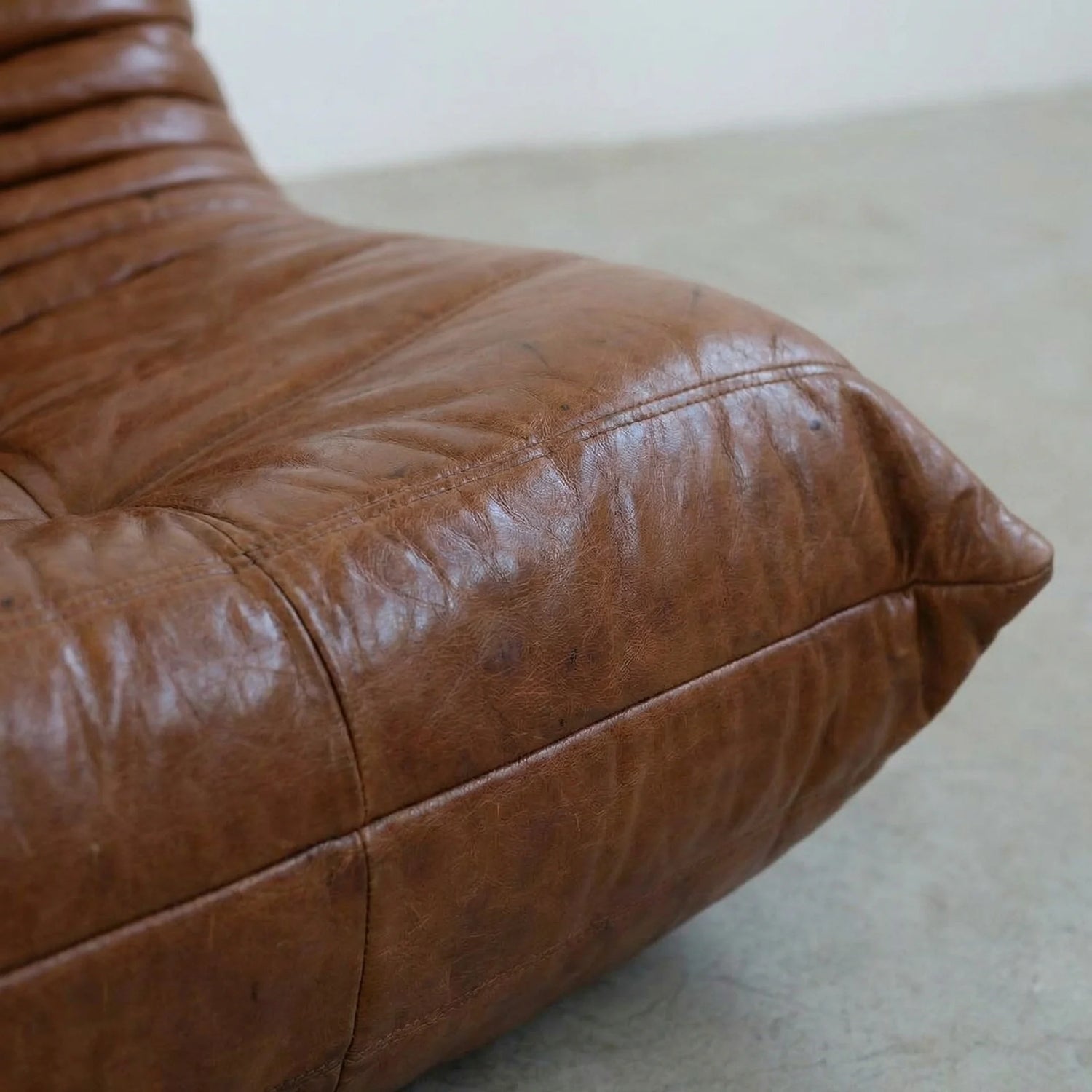 Close-up of the Togo Chair Dupe in brown real shiny oil wax leather on a concrete floor with a white wall background