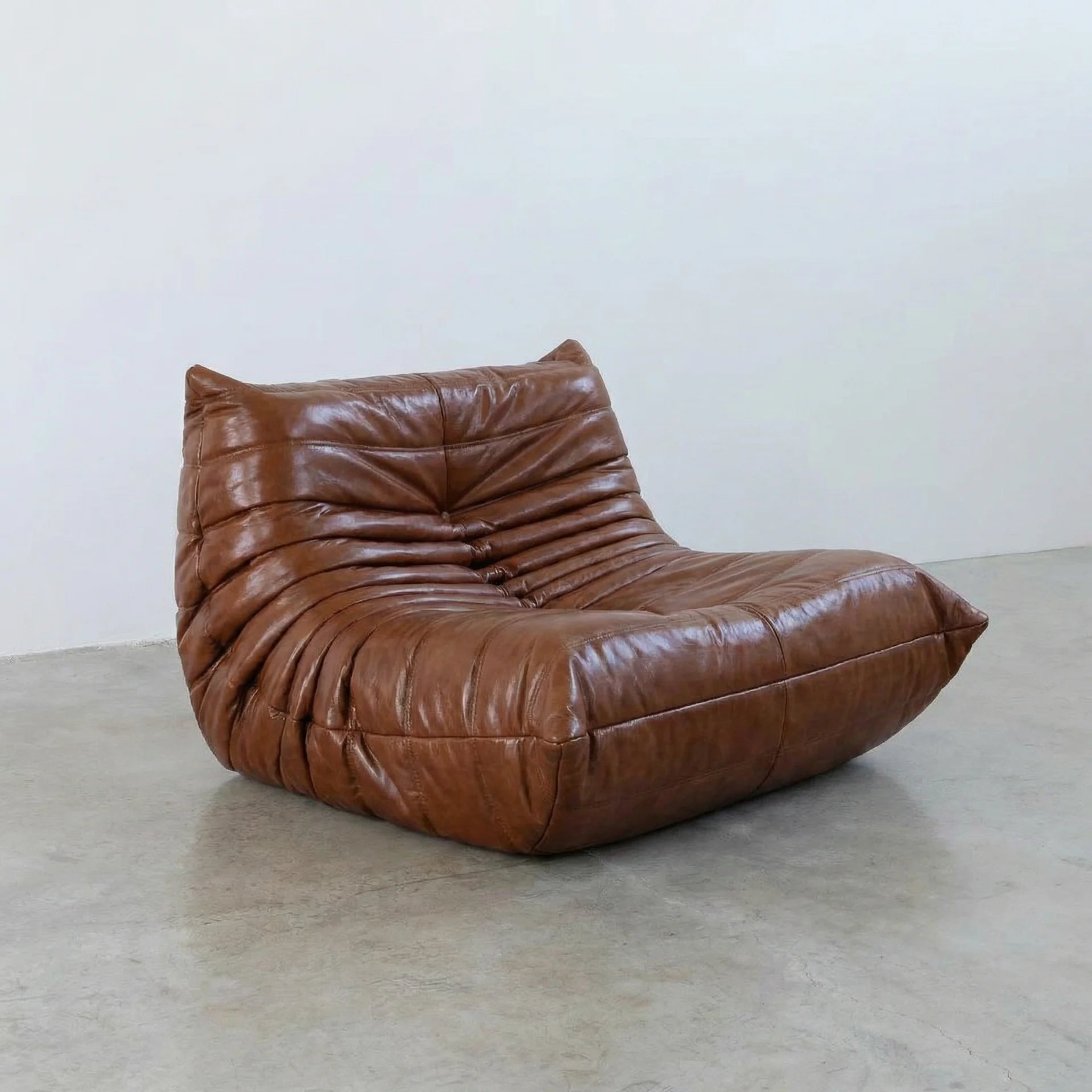 Togo Chair Dupe in brown real shiny oil wax leather on a concrete floor with a white wall background