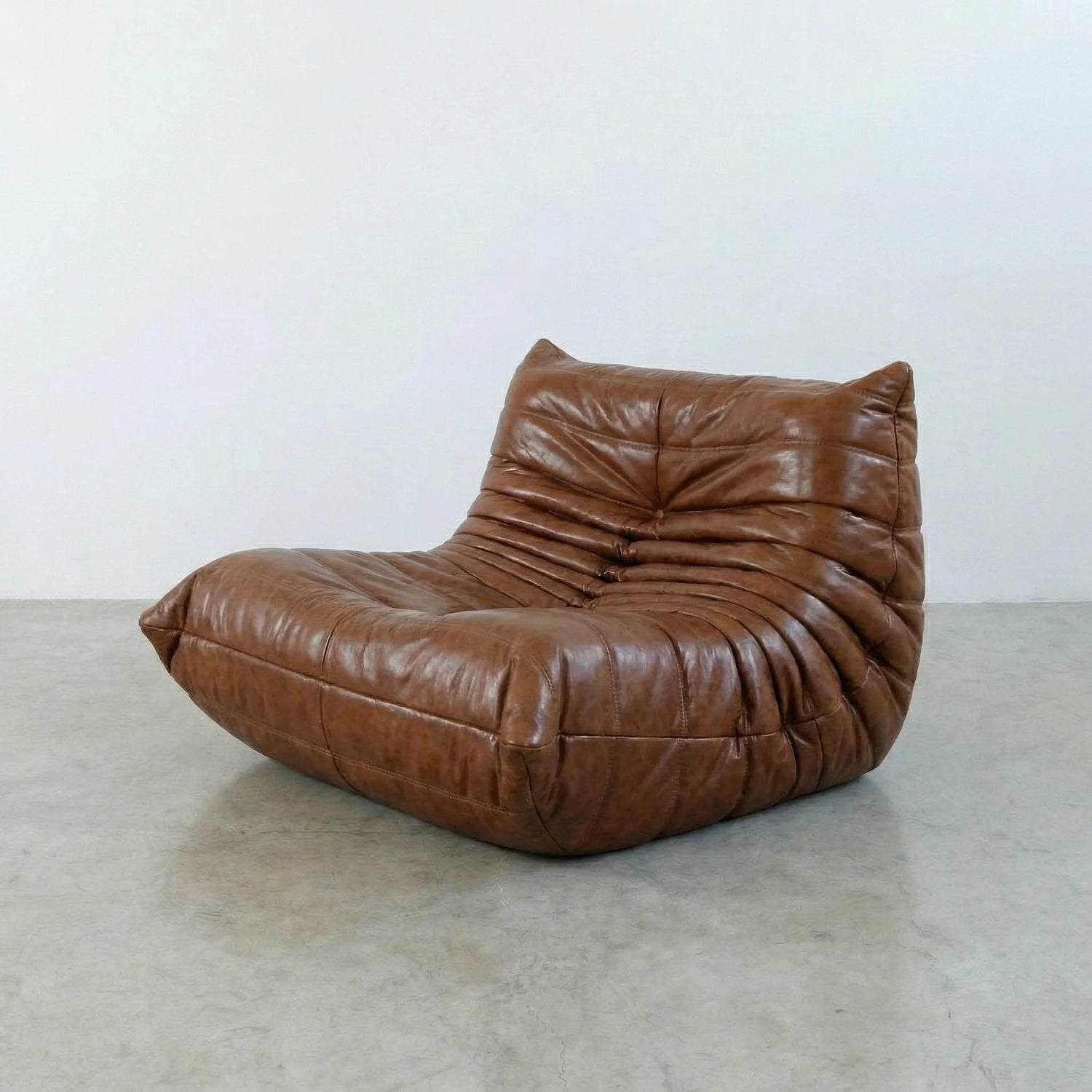 Togo Chair Dupe in brown real shiny oil wax leather on a concrete floor with a white wall background