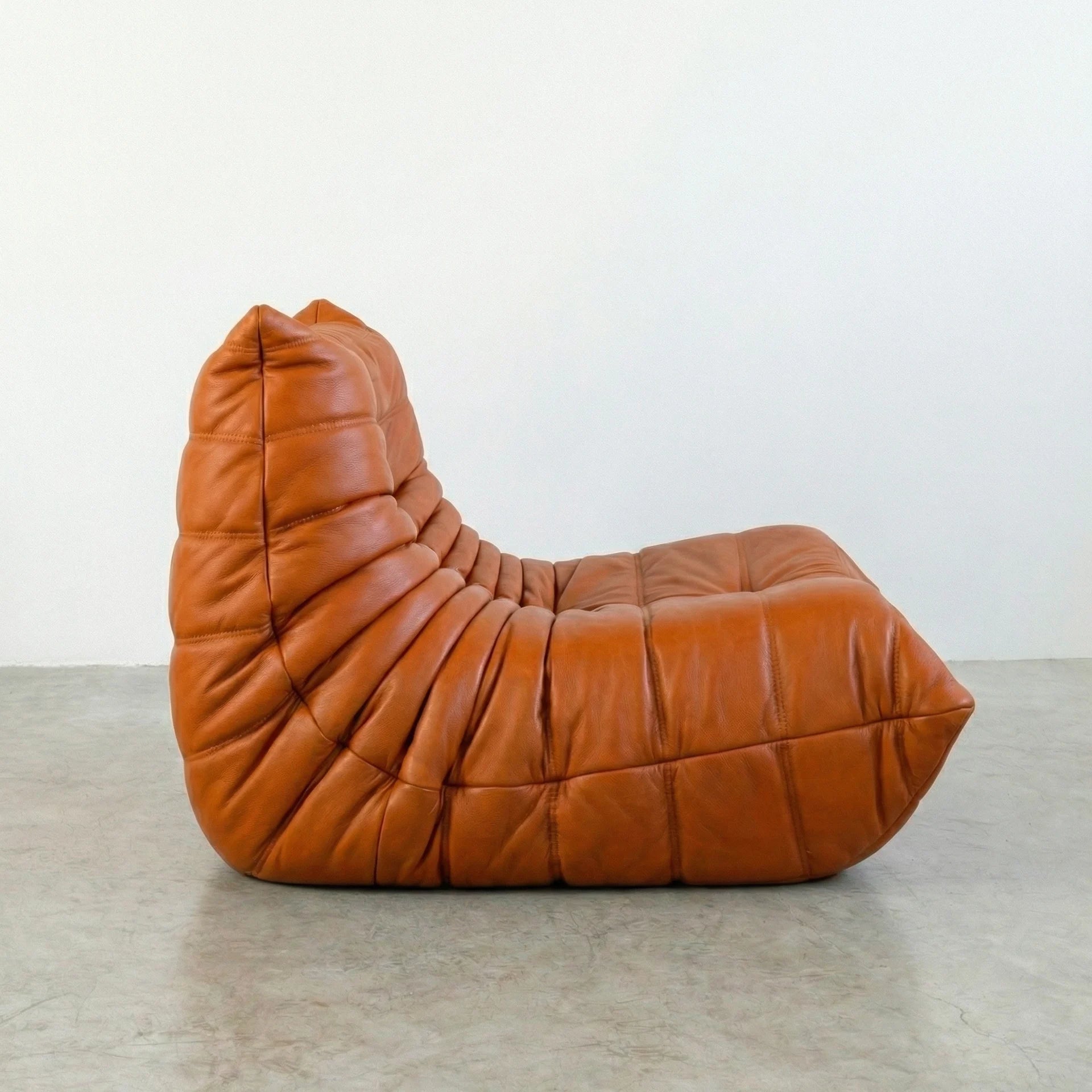 Togo Chair Dupe in cognac real cowhide leather on a concrete floor with a white wall background, shot from a side angle