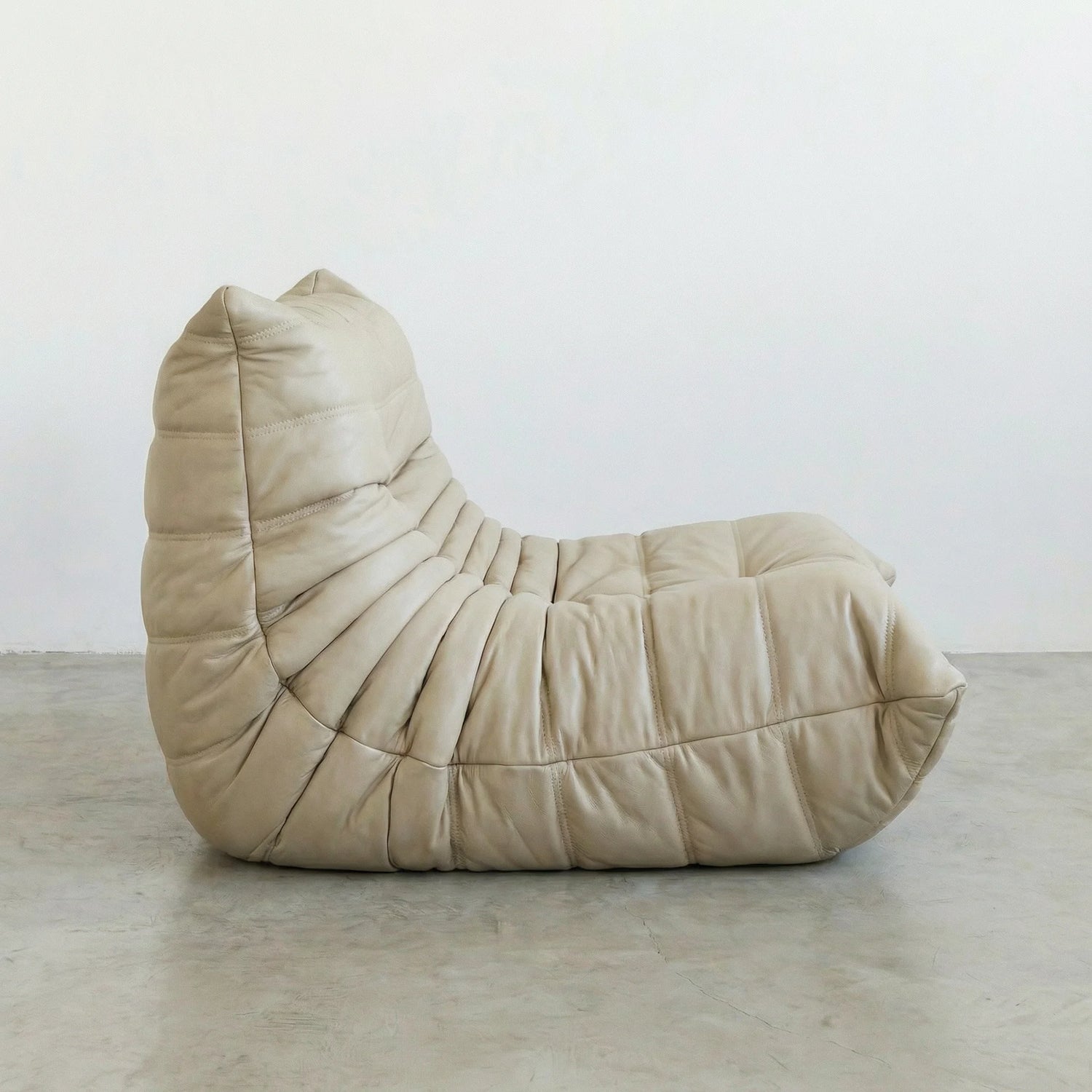 Togo Chair Dupe in cream real cowhide leather on a concrete floor with a white wall background, shot from a side angle