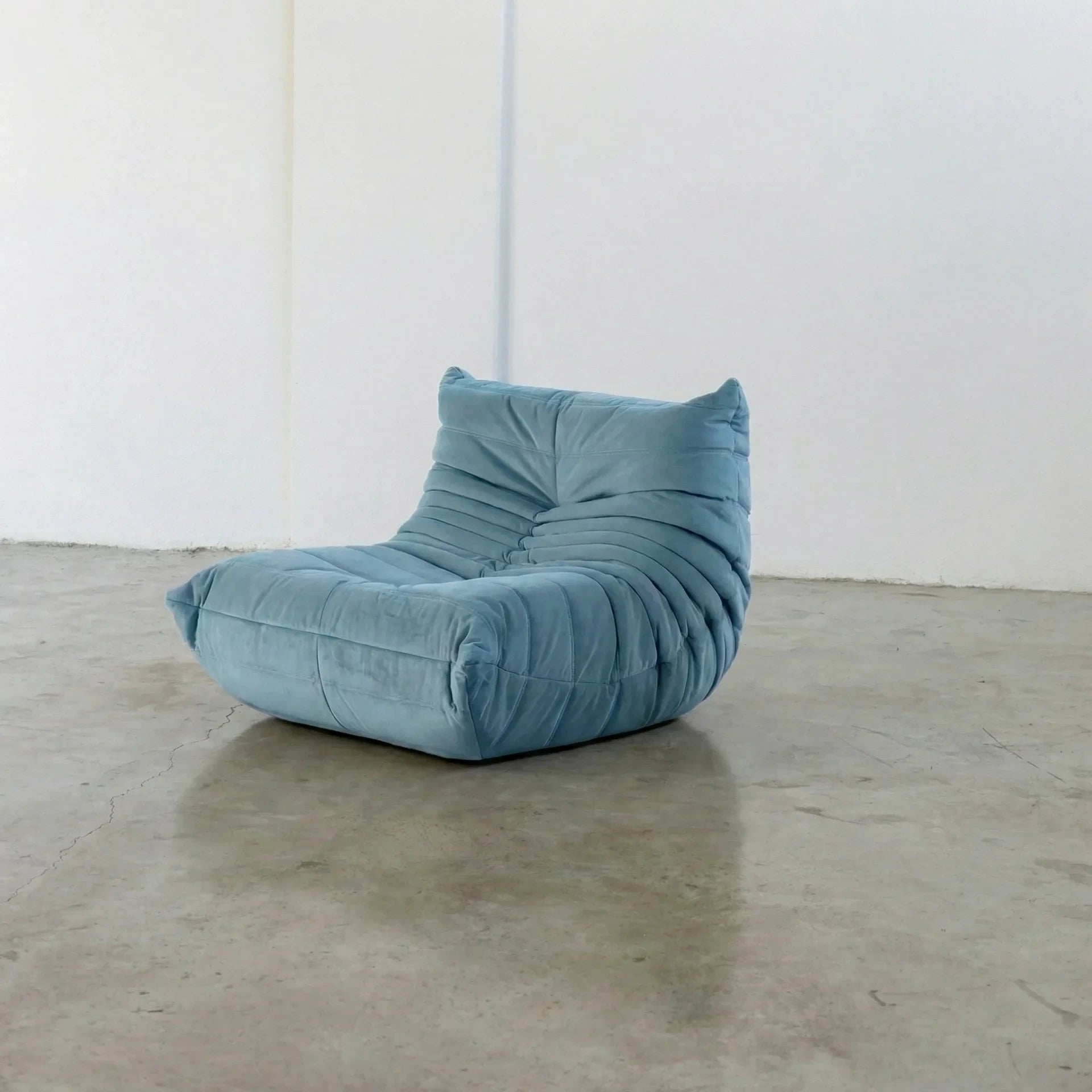 Handmade togo sofa chair as dupe in light blue velvet suede on a concrete floor with a white wall background