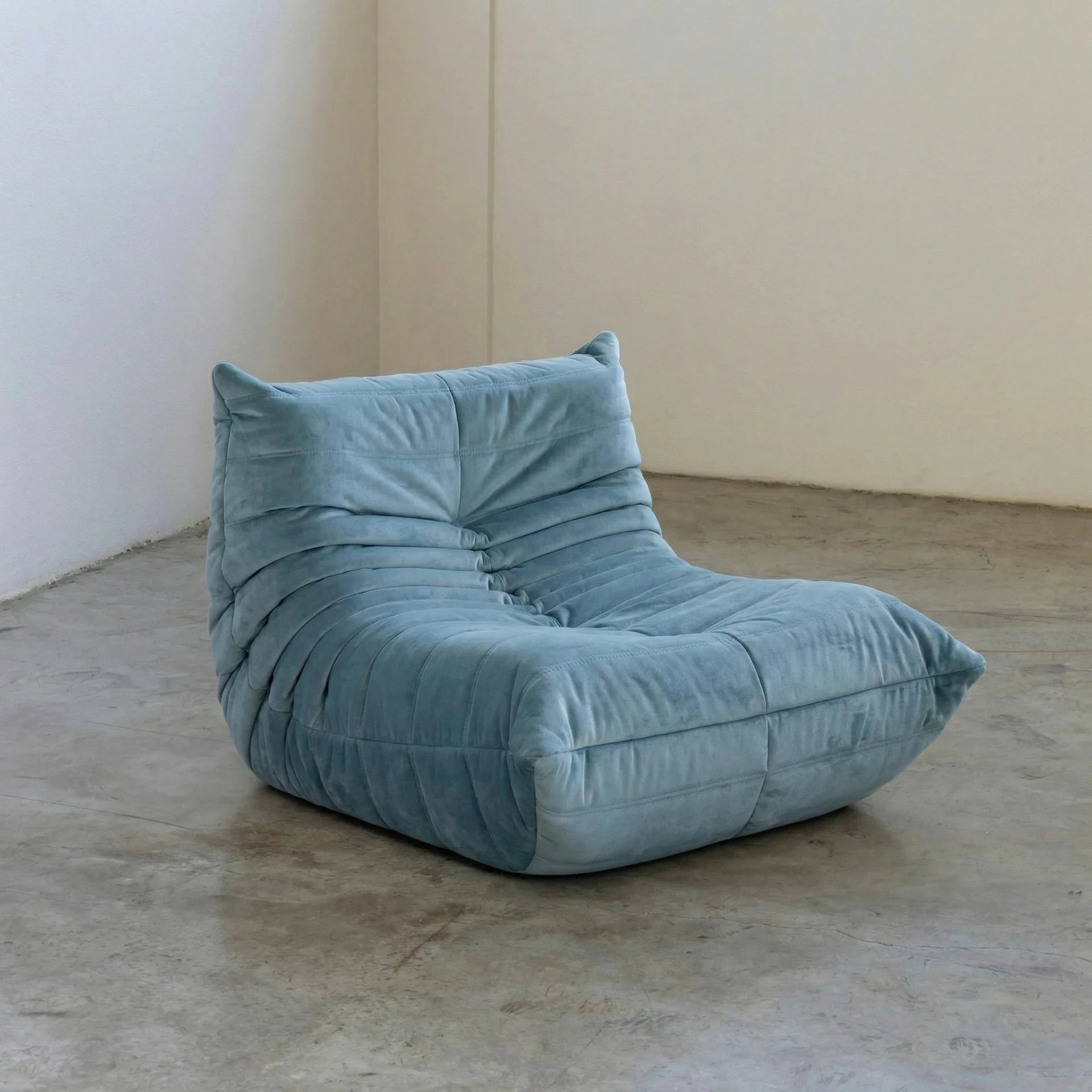 Handmade togo sofa chair as dupe in light blue velvet suede on a concrete floor with a white wall background
