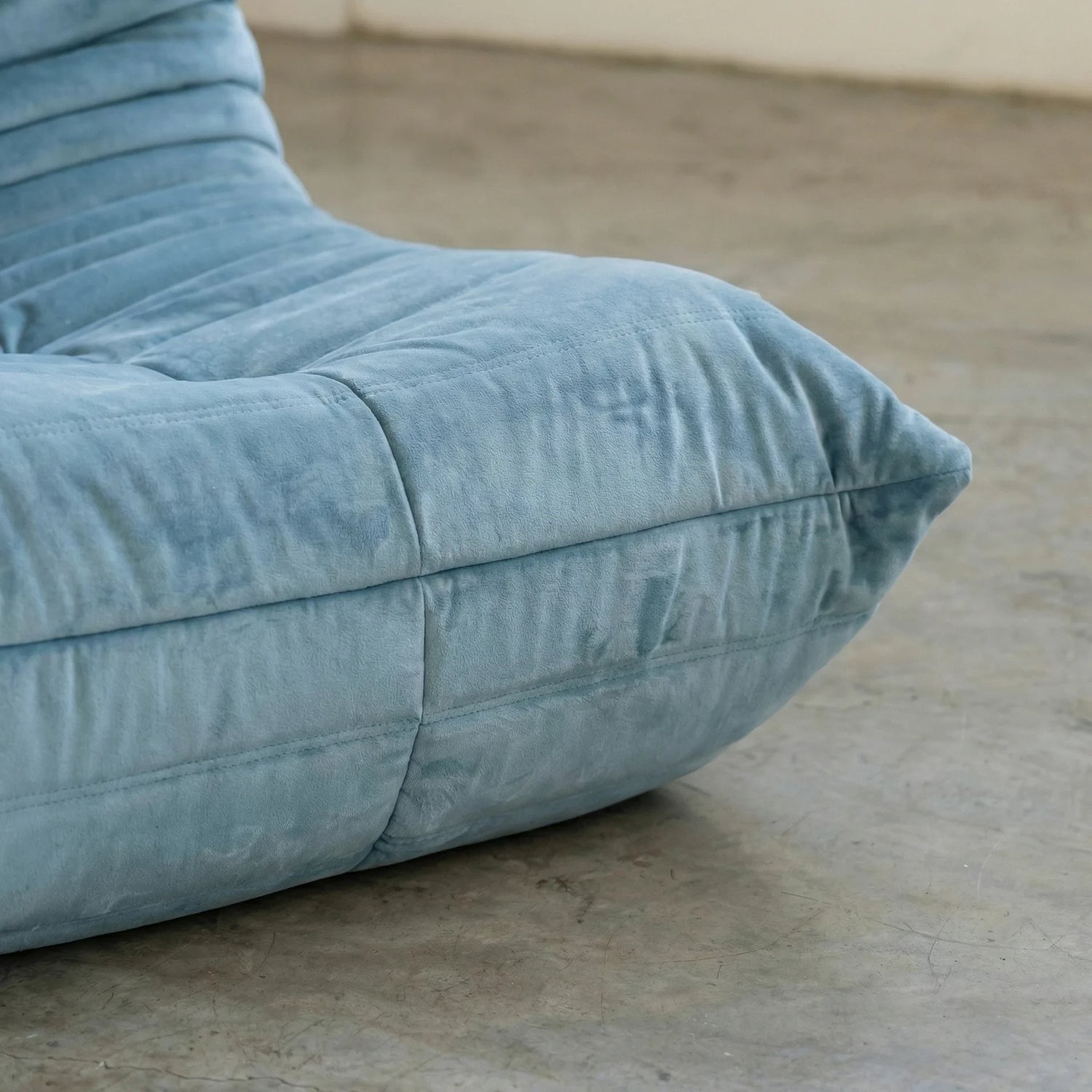 Close up of the handmade togo sofa chair as dupe in light blue velvet suede on a concrete floor with a white wall background