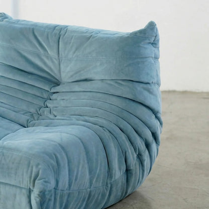 Close up of a handmade togo sofa chair as dupe in light blue velvet suede on a concrete floor with a white wall background