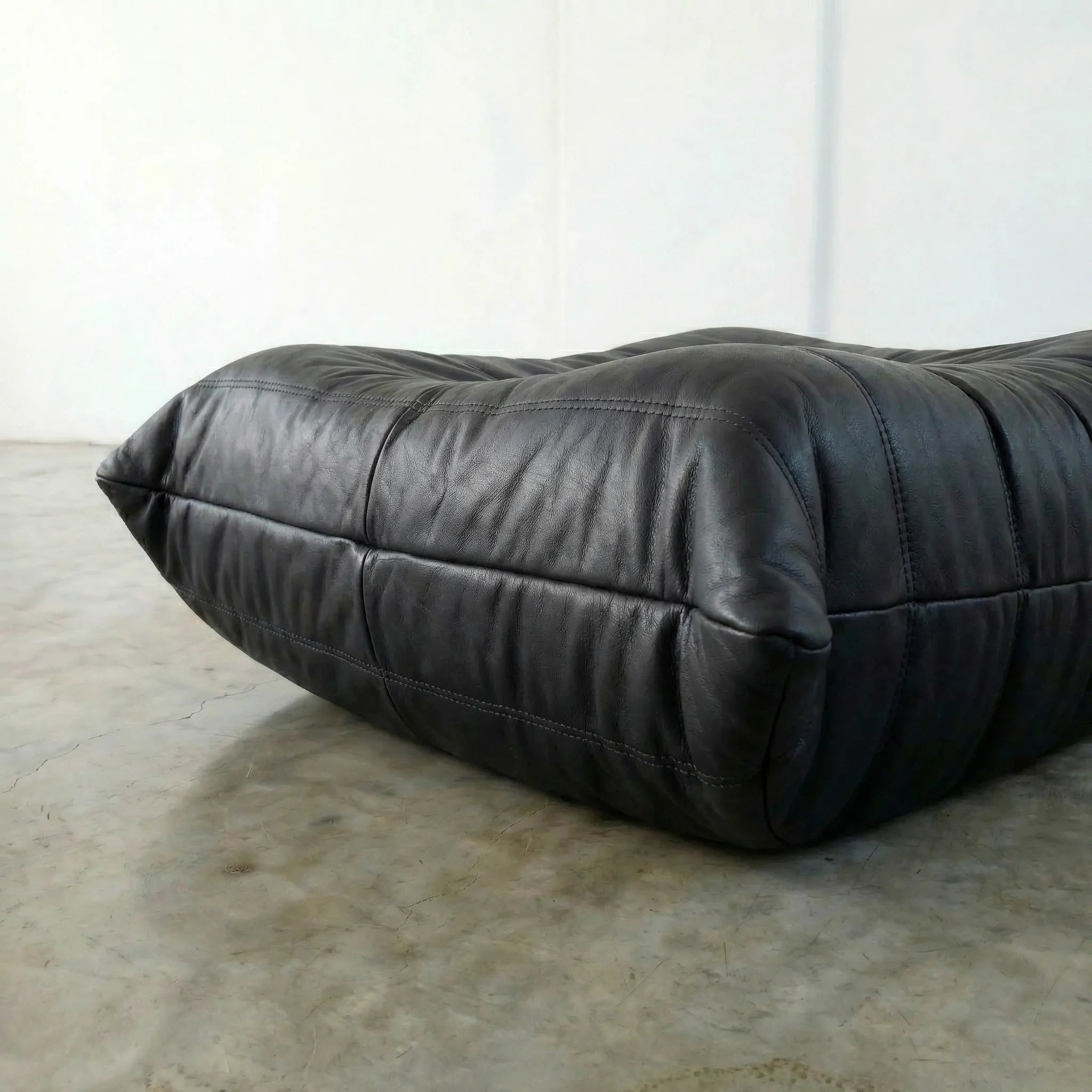 Black real leather togo pouf dupe on a concrete floor with a white wall background
