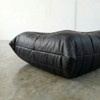 Black real leather togo pouf dupe on a concrete floor with a white wall background