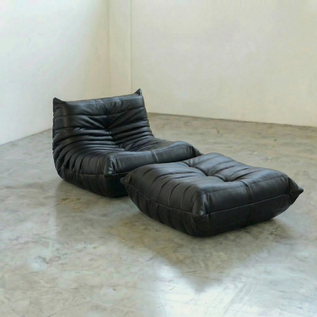 Black togo leather lounge chair and ottoman dupe on a concrete floor with a white wall background