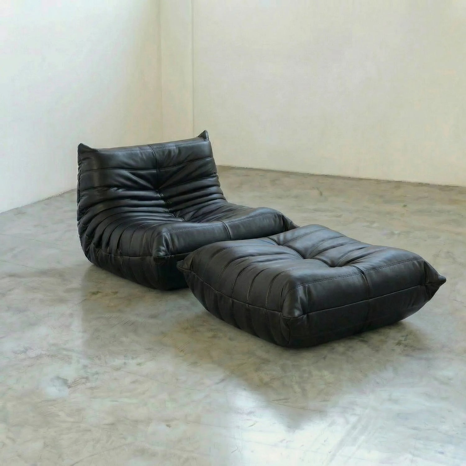Black togo leather lounge chair and ottoman dupe on a concrete floor with a white wall background