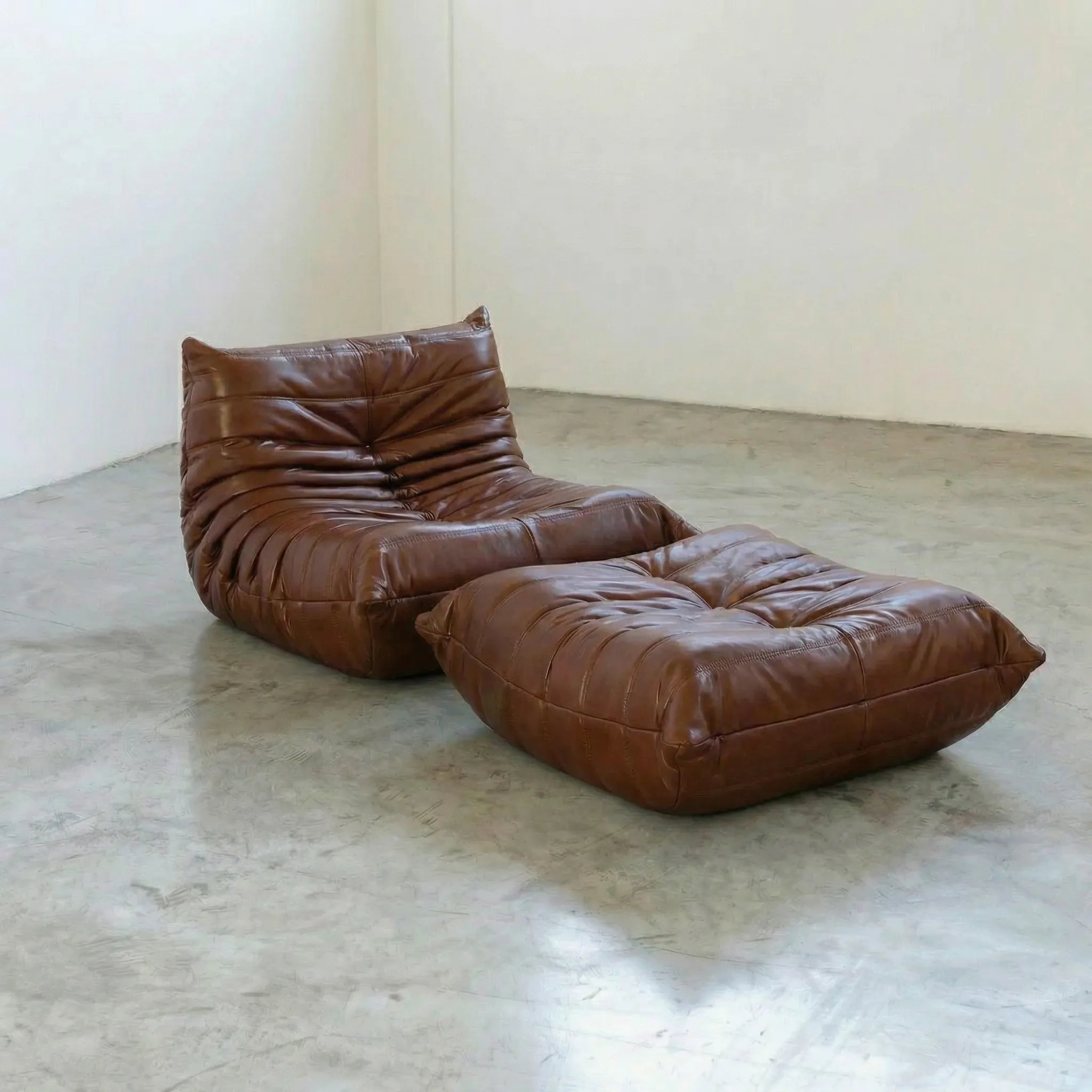 Brown togo dupe leather lounge chair and ottoman on a concrete floor with white walls.