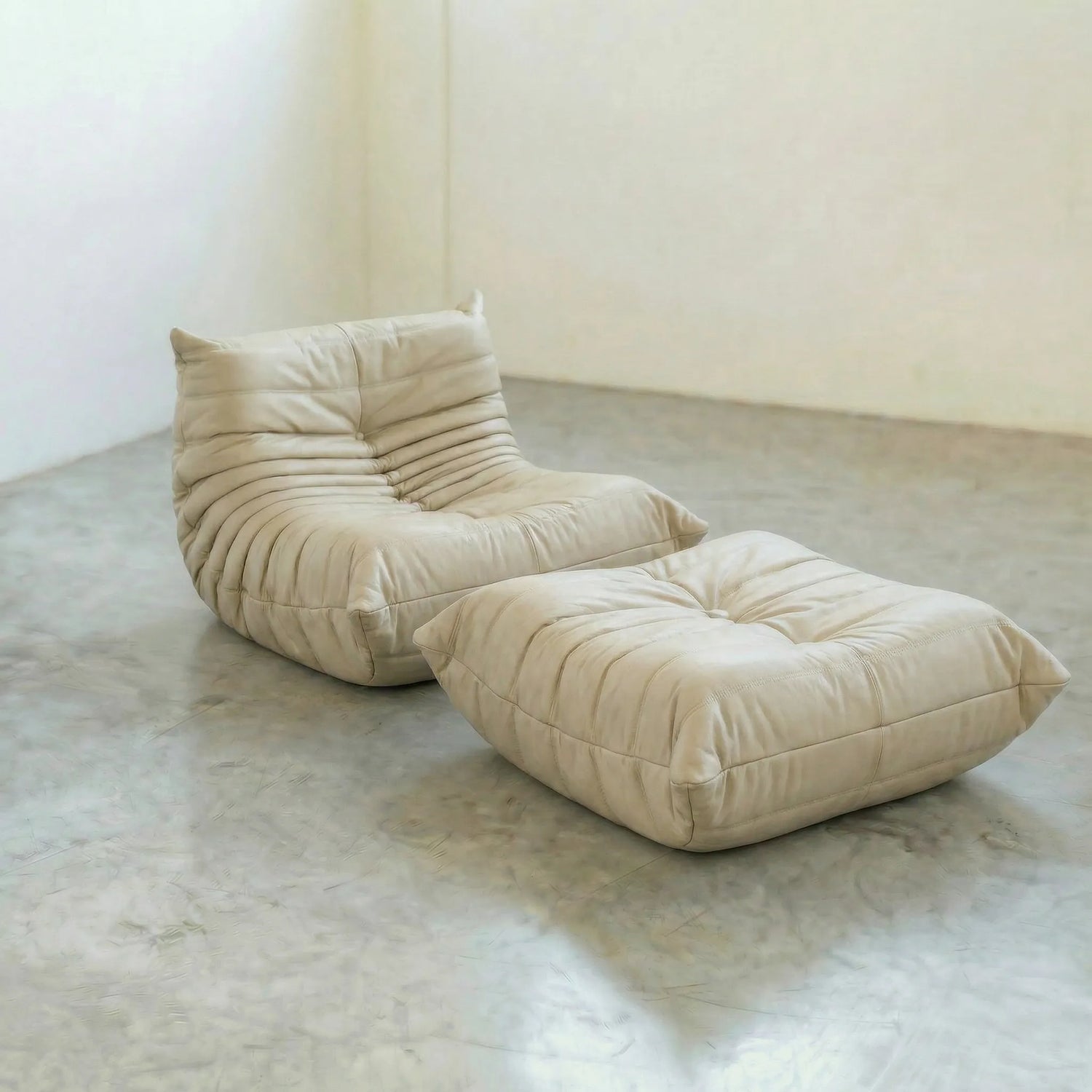Beige togo leather chair dupe and ottoman on a concrete floor with white walls.