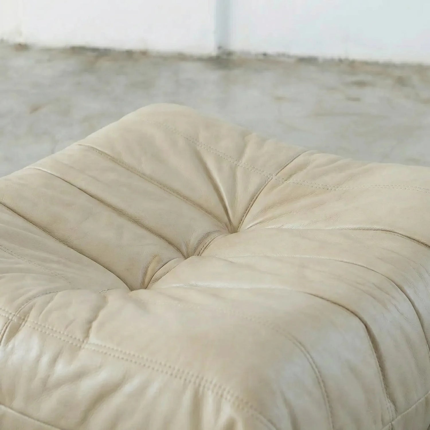 Beige togo leather ottoman dupe on a concrete floor with white walls