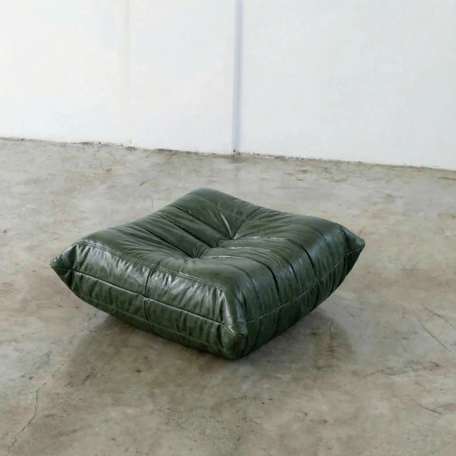 Green togo leather ottoman dupe on a concrete floor with white walls.