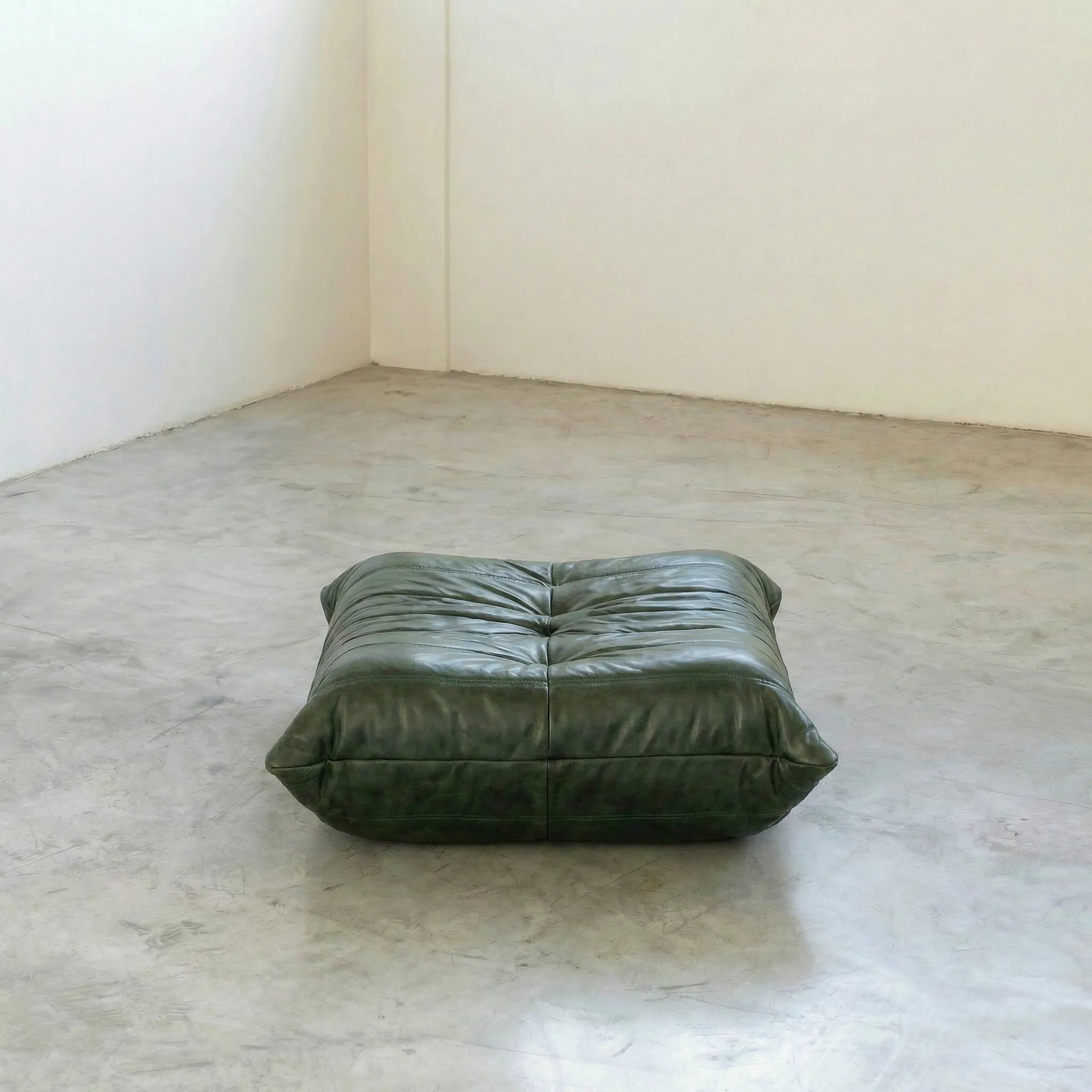 Green togo leather ottoman dupe on a concrete floor with white walls.
