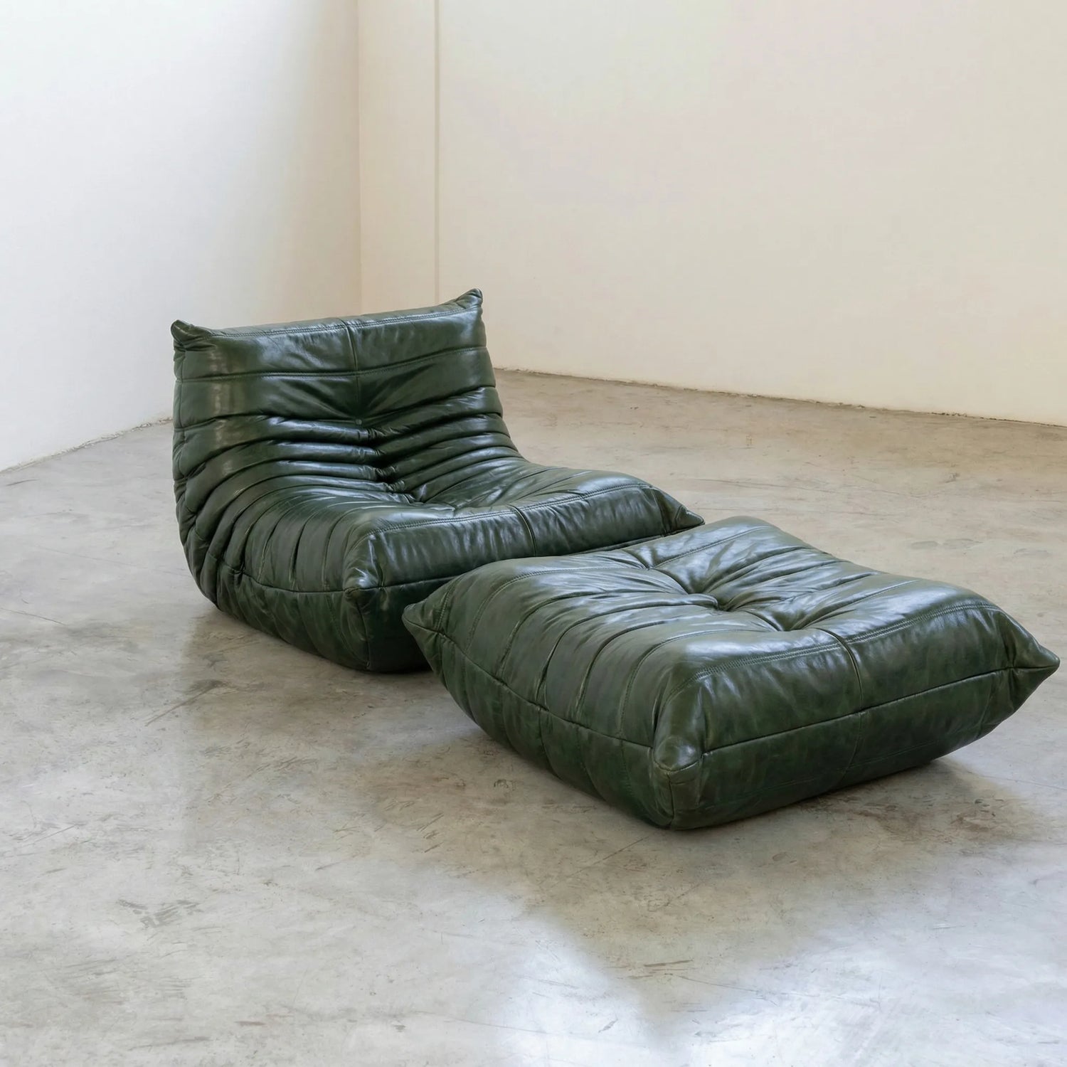 Green togo leather lounge chair dupe and ottoman on a concrete floor with white walls.