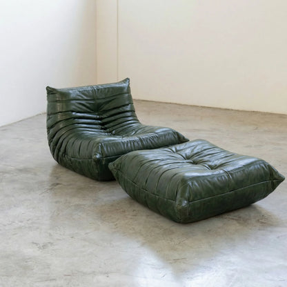 Green togo leather lounge chair dupe and ottoman on a concrete floor with white walls.
