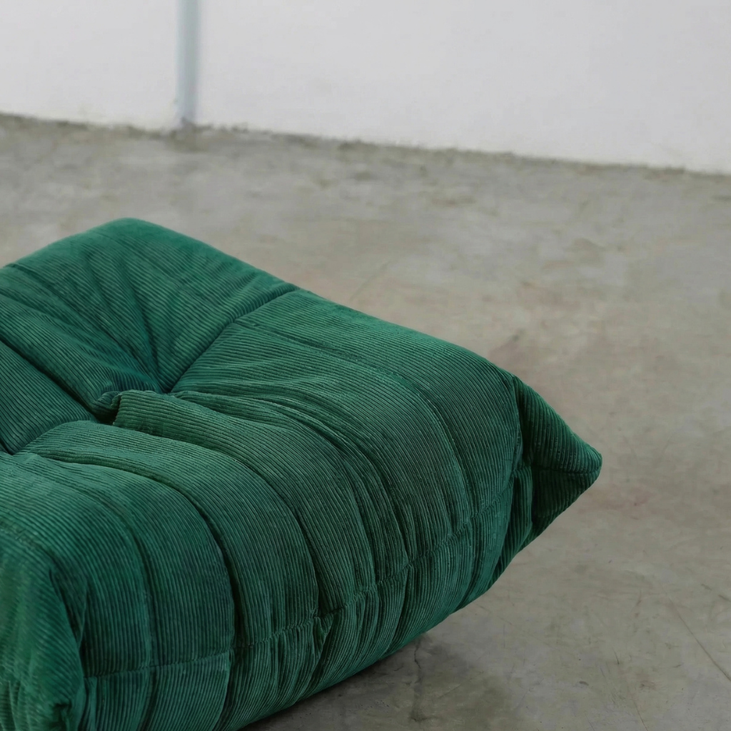 Close-up of dark green togo ottoman replica in corduroy on a concrete floor.