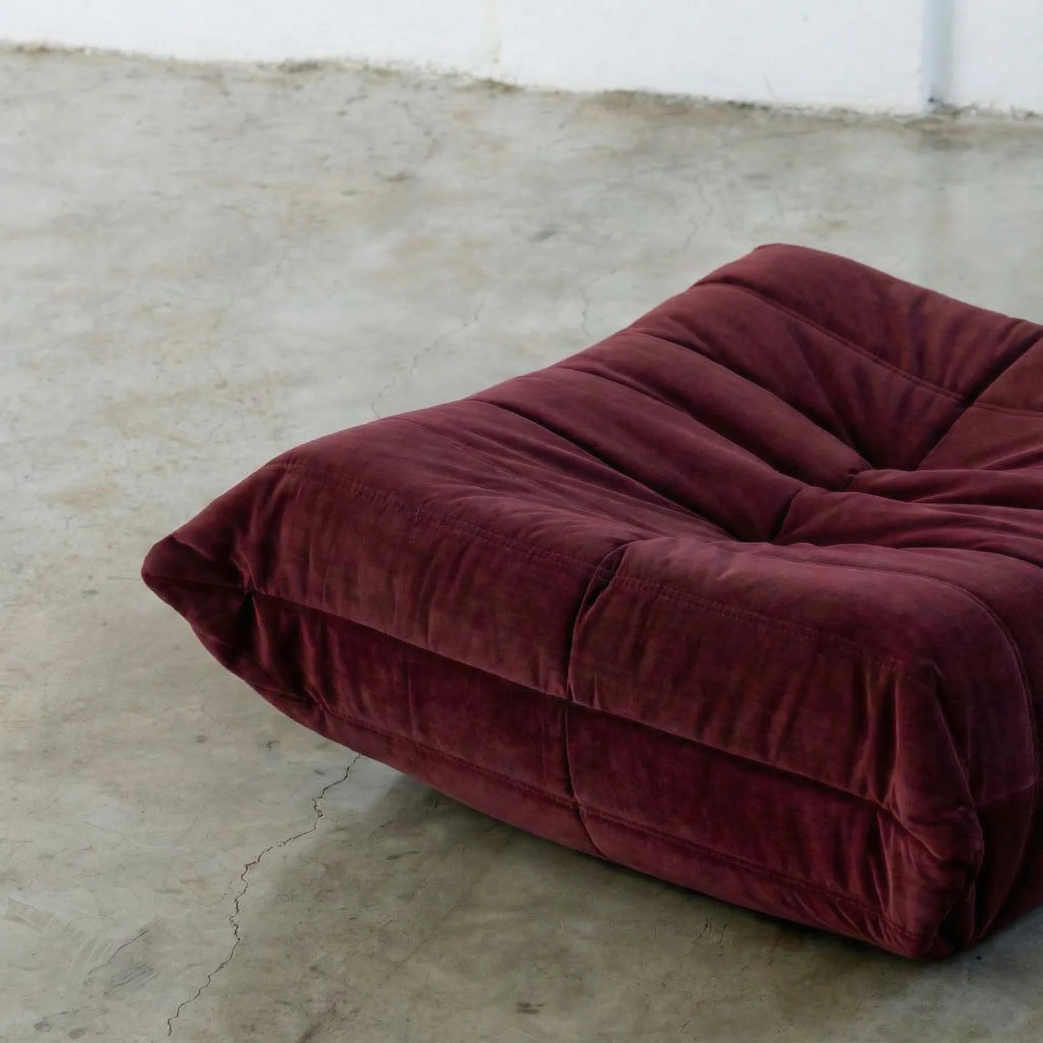 Close-up of burgundy togo ottoman replica in velvet suede on a concrete floor.
