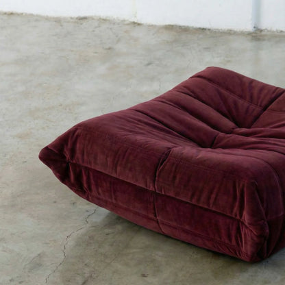 Close-up of burgundy togo ottoman replica in velvet suede on a concrete floor.