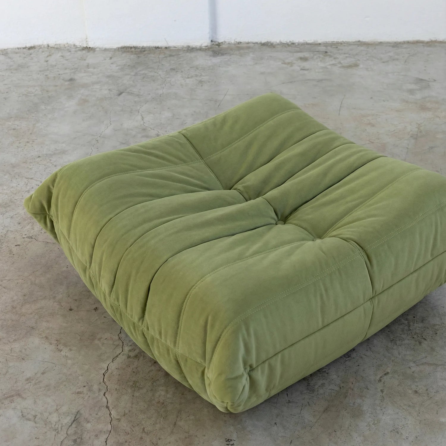Close-up of green togo ottoman replica in velvet suede on a concrete floor.