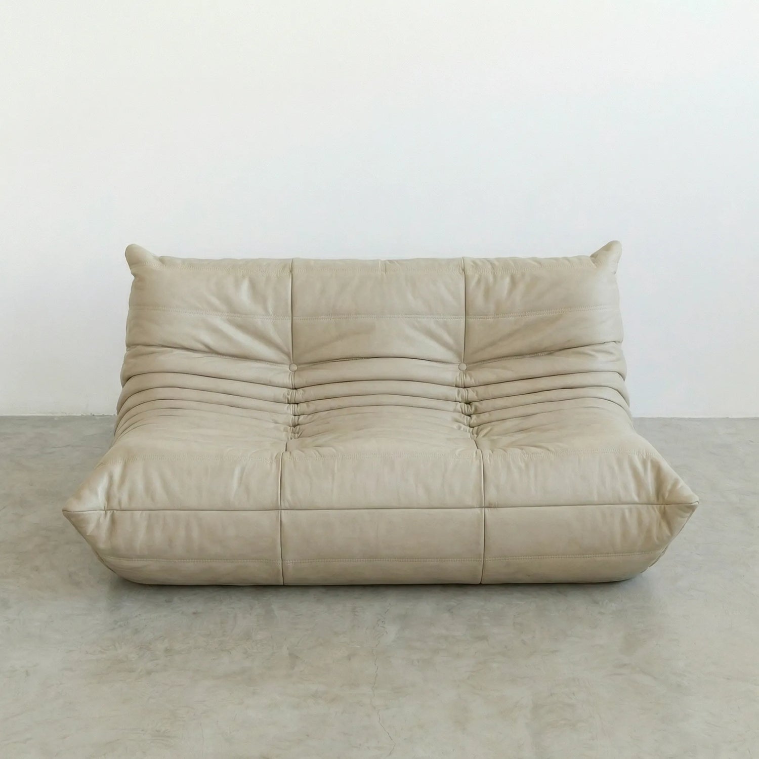 Beige togo sofa dupe in leather on a concrete floor with a white wall background
