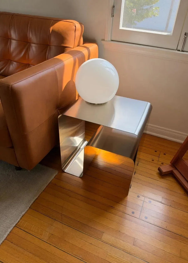 Reflective curved steel side table with a white globe lamp beside a brown sofa.