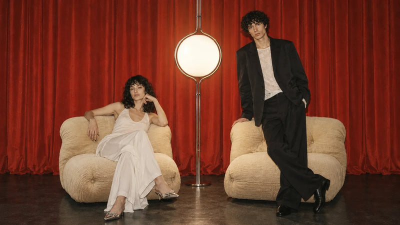 Two people beside cream lounge chairs and a globe lamp against red curtains.