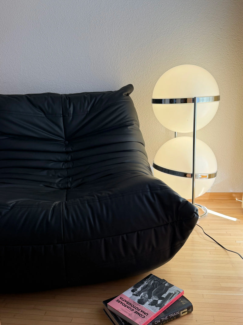 A modern black leather lounge chair in a minimalist room next to a zuru two-tier spherical floor lamp with chrome accents, with a book on a light wood floor in the foreground.